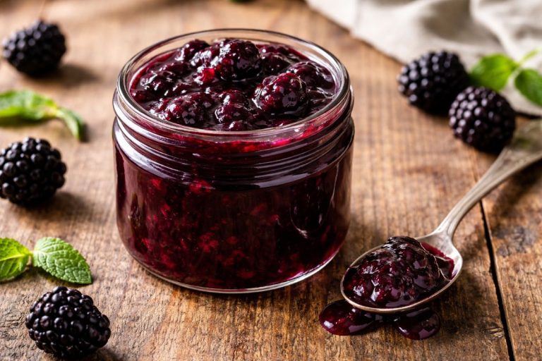 Thick glossy blackberry compote in a glass jar with visible berry pieces on a rustic wooden table with a spoon beside it